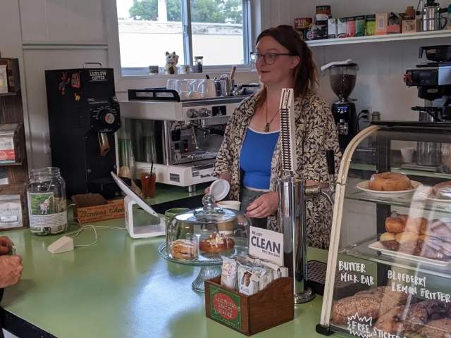 A woman stands behind the green counter. A display of pastries is on the right.