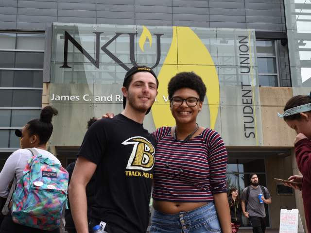 Two students face the camera in front of the student union sign. Other students are walking behind or next, not looking at the camera.