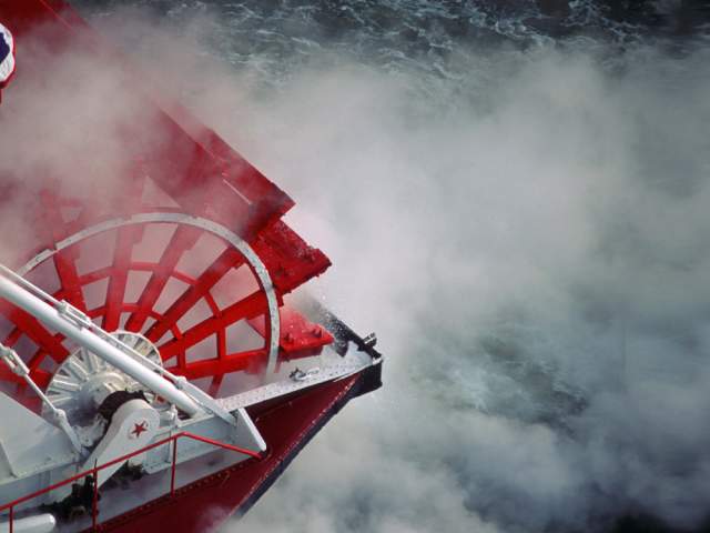 A red paddlewheel on a red and white boat. Steam and mist churn up around the wheel.