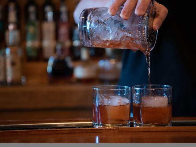 A close up of two drinks being poured over large, square ice in rocks glasses.