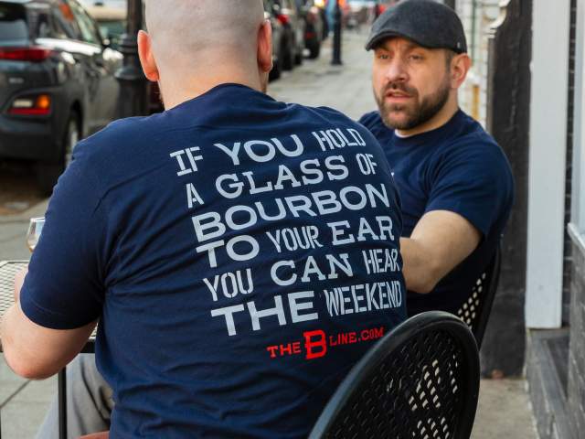 A man sits facing away from the camera. The back of his shirt reads "If you hold a glass of bourbon to your ear you can hear the weekend". Behind him is another man, facing the camera and looking at the first man.