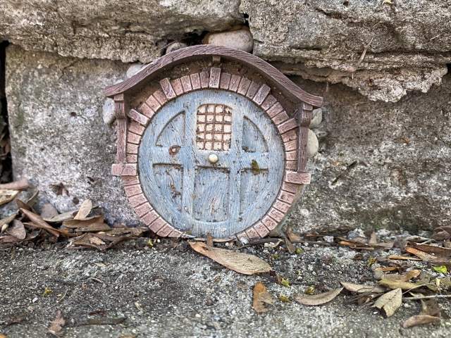 Image is of a small, round, blue fairy door that is along the side walk, built into the retaining wall.