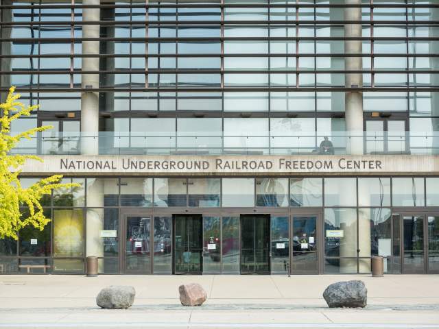 The glass walled front entrance of the National Underground Railroad Freedom Center with a tree, a man, and large stones.
