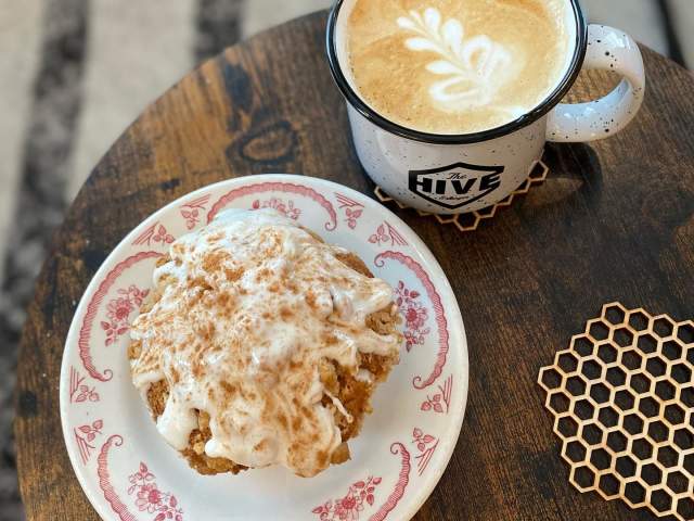 The image is an ariel view of a saucer plate with a muffin covered in icing next to a filled coffee mug on a wood table.