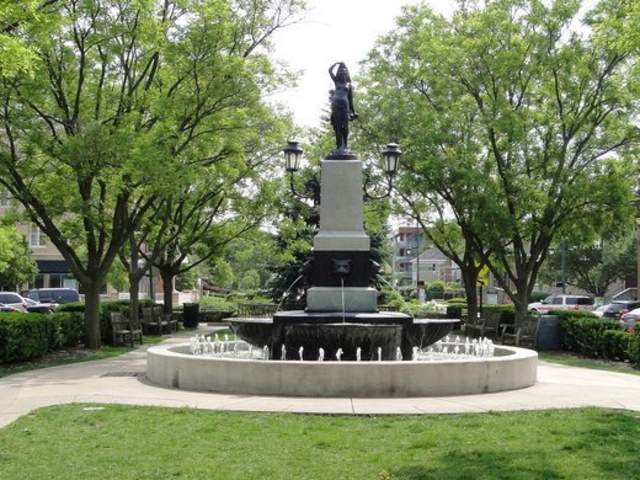 Image is of the Hyde Park statue that has water shooting out into a pool in the center of a town that splits the road with buildings on each side.