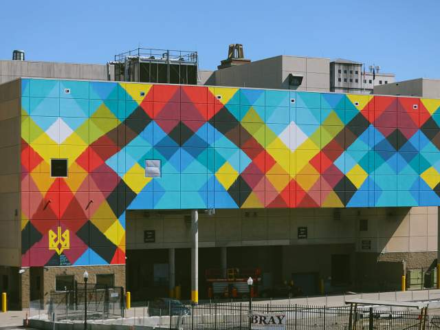 A large-scale mural on the side of a convention center features colorful criss-crossing line reminiscent of a weaving