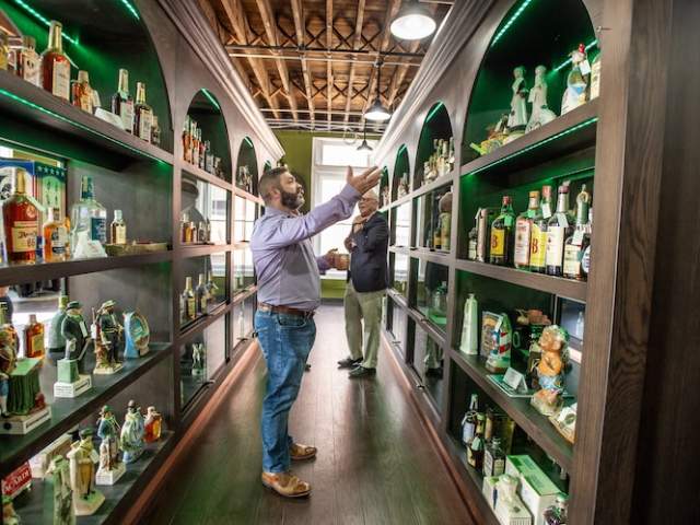 Two men stand looking at bookshelves filled with bottles of vintage spirits
