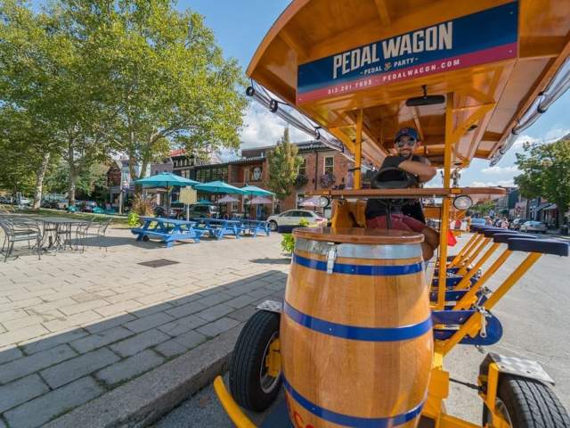 The front of the Covington Pedal Wagon with a smiling driver and Mainstrasse Village in the background on a sunny day
