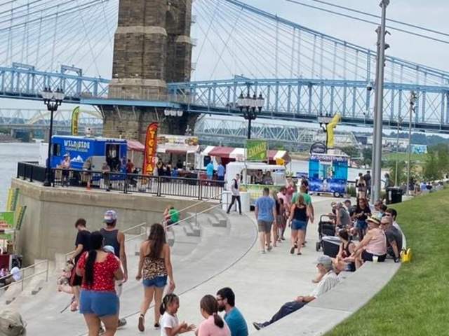 People mingle at a festival. In the background are several food trucks in front of the Roebling Suspension Bridge.