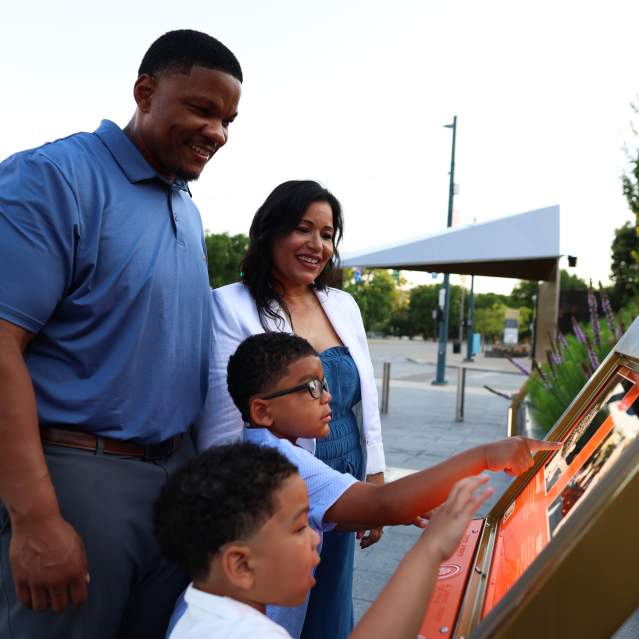A family interacts with an outdoor display at the Black Music Walk of Fame in the foreground, while a park with trees and a covered structure is visible in the background.