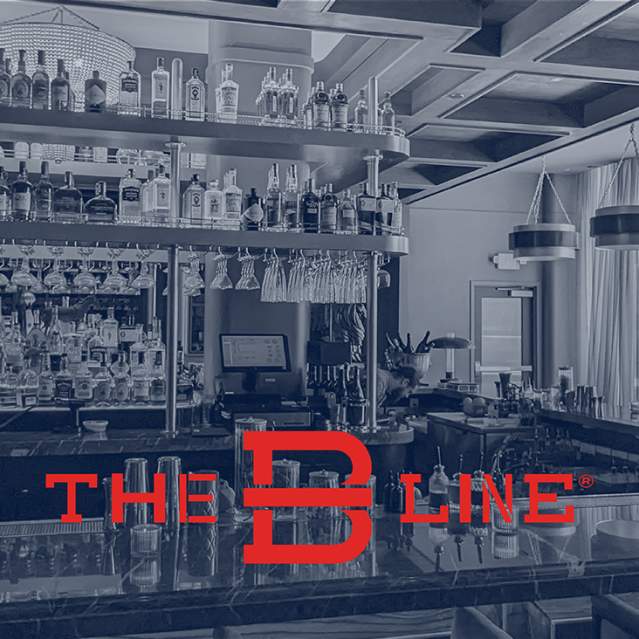 A sleek bar counter with glassware sits in the foreground, while shelves of liquor bottles and modern pendant lights fill the background. Text says "The B-Line".