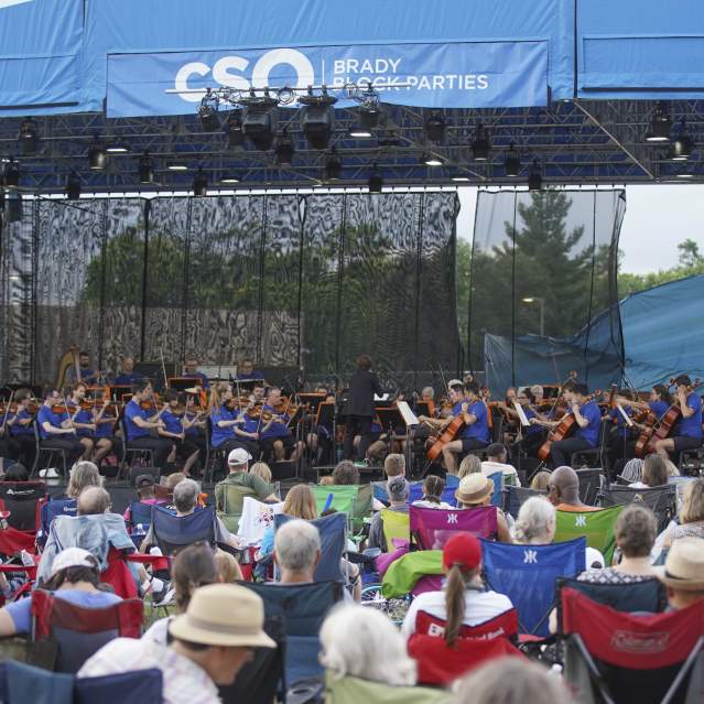 A crowd of people in pop-up lawn chairs sit by a stage and watch the Cincinnati Symphony Orchestra perform