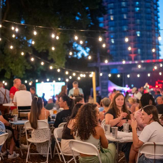 People are gathered at tables under string lights in the foreground, with a stage and a tall building visible in the background.
