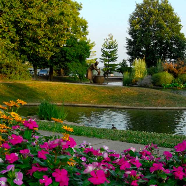 A walking path, flanked by flowers, runs next to a pond. Trees and a statue are in the background.