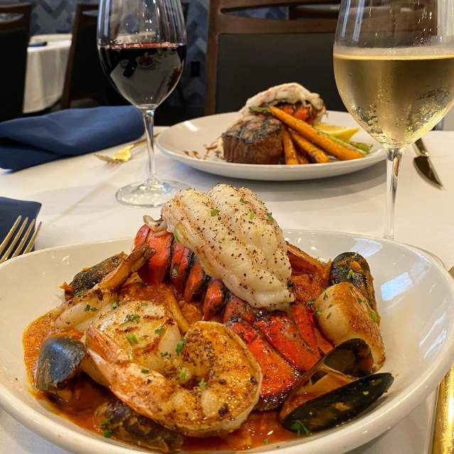 A close-up of a seafood platter with lobster, shrimp, and scallops in the foreground is set against a blurred background of a steak dinner and wine glasses on a white tablecloth.