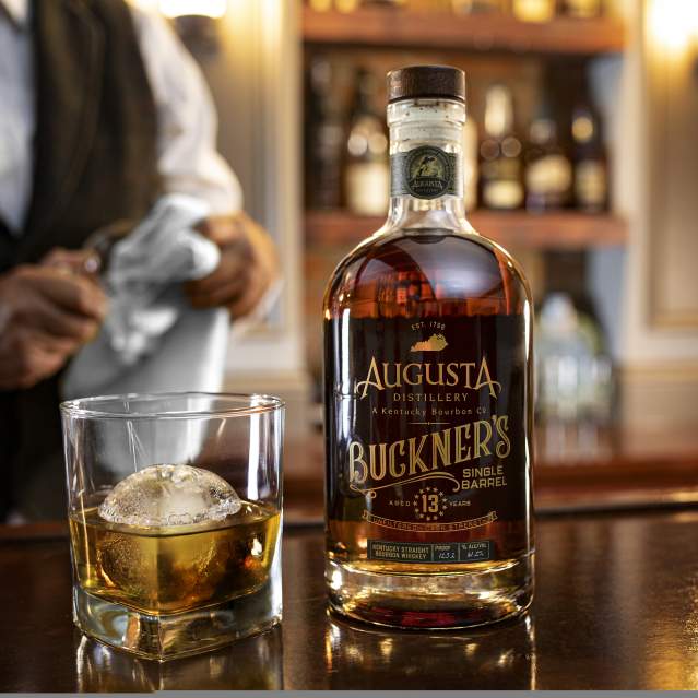 Image is of a bottle of Augusta Distillery's Buckner's Single Barrell and a glass of bourbon next to it with an ice ball.
