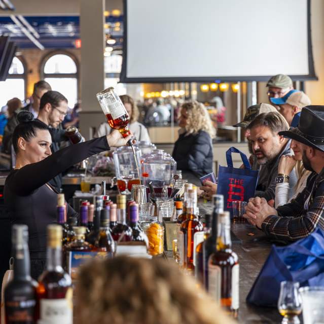 An array of bottles line the bar while a bartender pours bourbon into a glass.