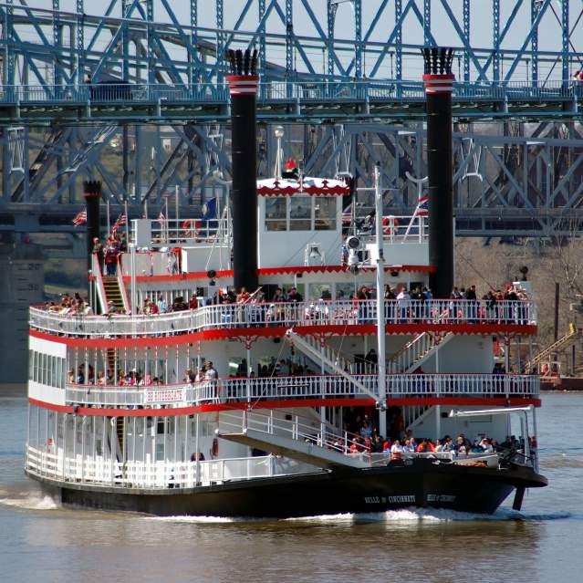 A red and white, paddlewheel riverboat cruises on the water. There are 3 decks with people standing on them.