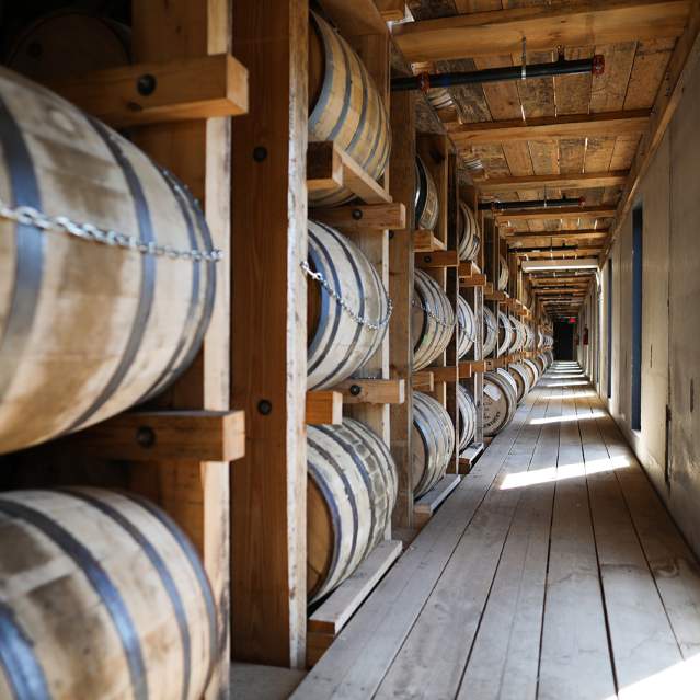 Rows of bourbon barrels line the left side of the photo in a wooden rickhouse.