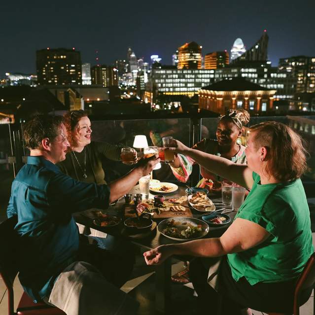 Four people toast each other at night with the Cincinnati skyline in the background