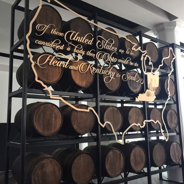 A rack of bourbon barrels with a gold, stenciled map of the U.S.and the words "If the United States are to be considered a body then Ohio would be its heart and Kentucky its soul.