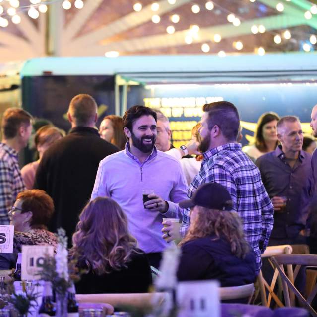 A group of people stands around under a tent with string lights drinking beer