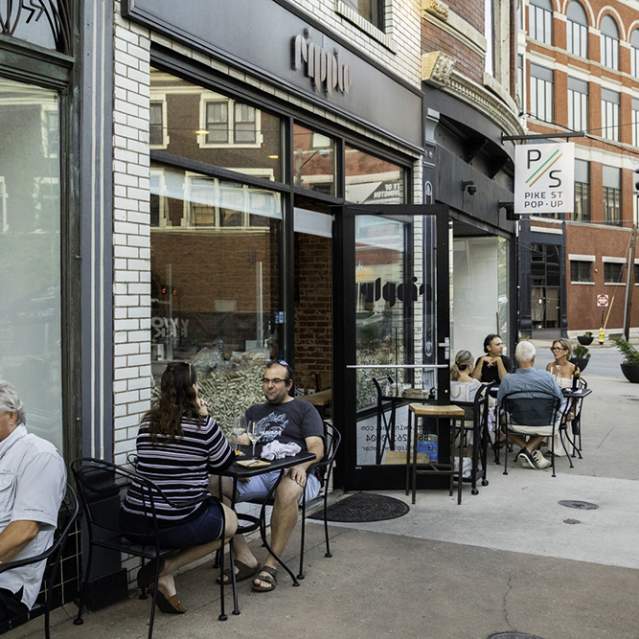 A group of people sits at black metal patio tables on a sidewalk outside Ripple Wine Bar
