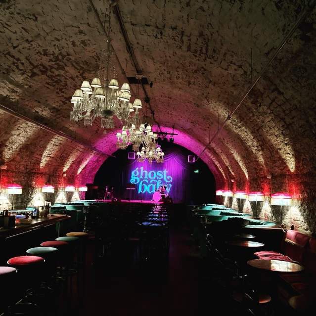 A tunnel lit with white and pink lights with a sign at the end in blue reading ghost baby. Rows of barstools line the walls of the tunnel
