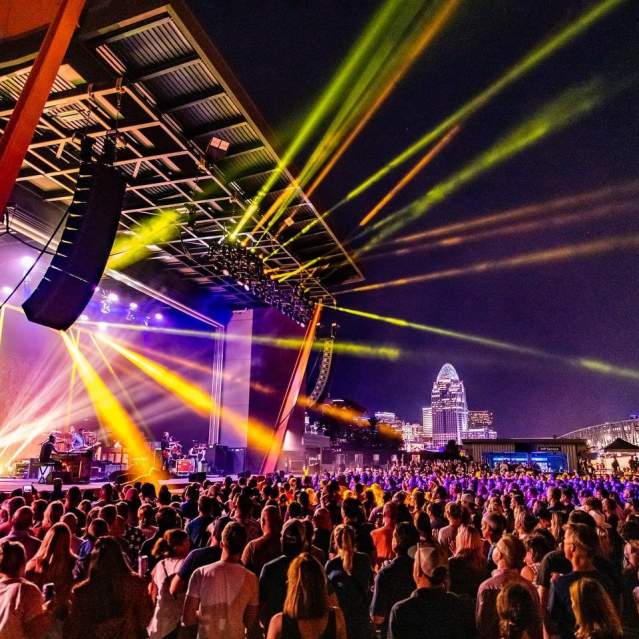 Night concert outdoors with rays of yellow light at Promowest Pavilion in Newport, Ky, with the Cincinnati skyline in the background
