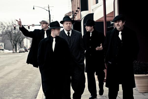 A group of men walk towards the camera dressed in stereotypical mob attire.