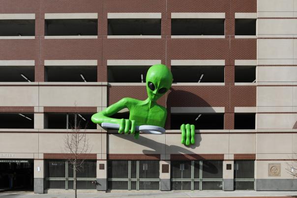 A green alien leans out of a parking garage, looking over the sidewalk with a magnifying glass.