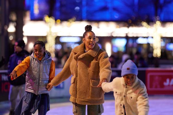 A mom and her two children ice skate while holding hands.