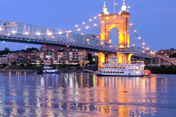 The Roebling Suspension bridge connecting Northern Kentucky to Cincinnati, with the Ohio river looking pink and blue in the sunset and a BB Riverboat sailing under the bridge