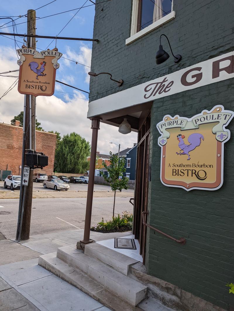 The corner of a green brick building with two yellow signs that both have a purple rooster painted on them and the words, "Purple Poulet: A Southern Bistro"