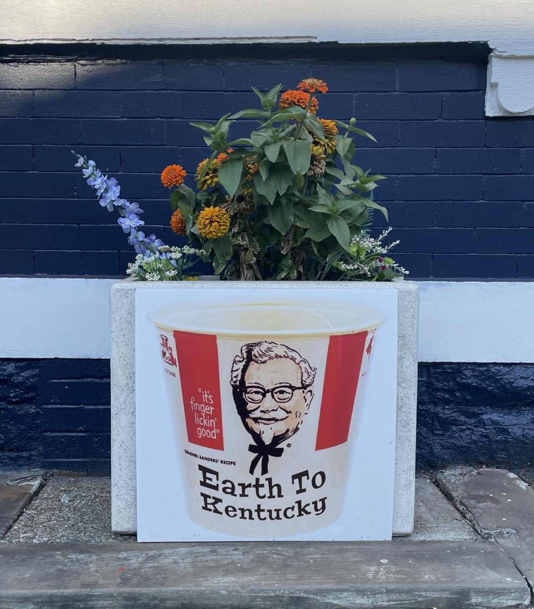 Flower pot on the outside of the building with an image of the KFC bucket of chicken with Sanders on it.