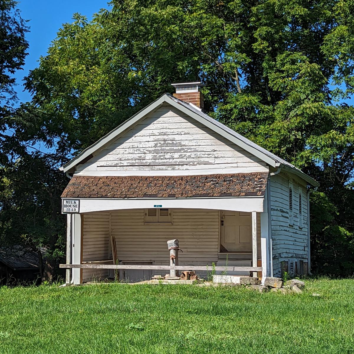 Image is of the Milk House, it is a white, one room, one story building with the door on the right.
