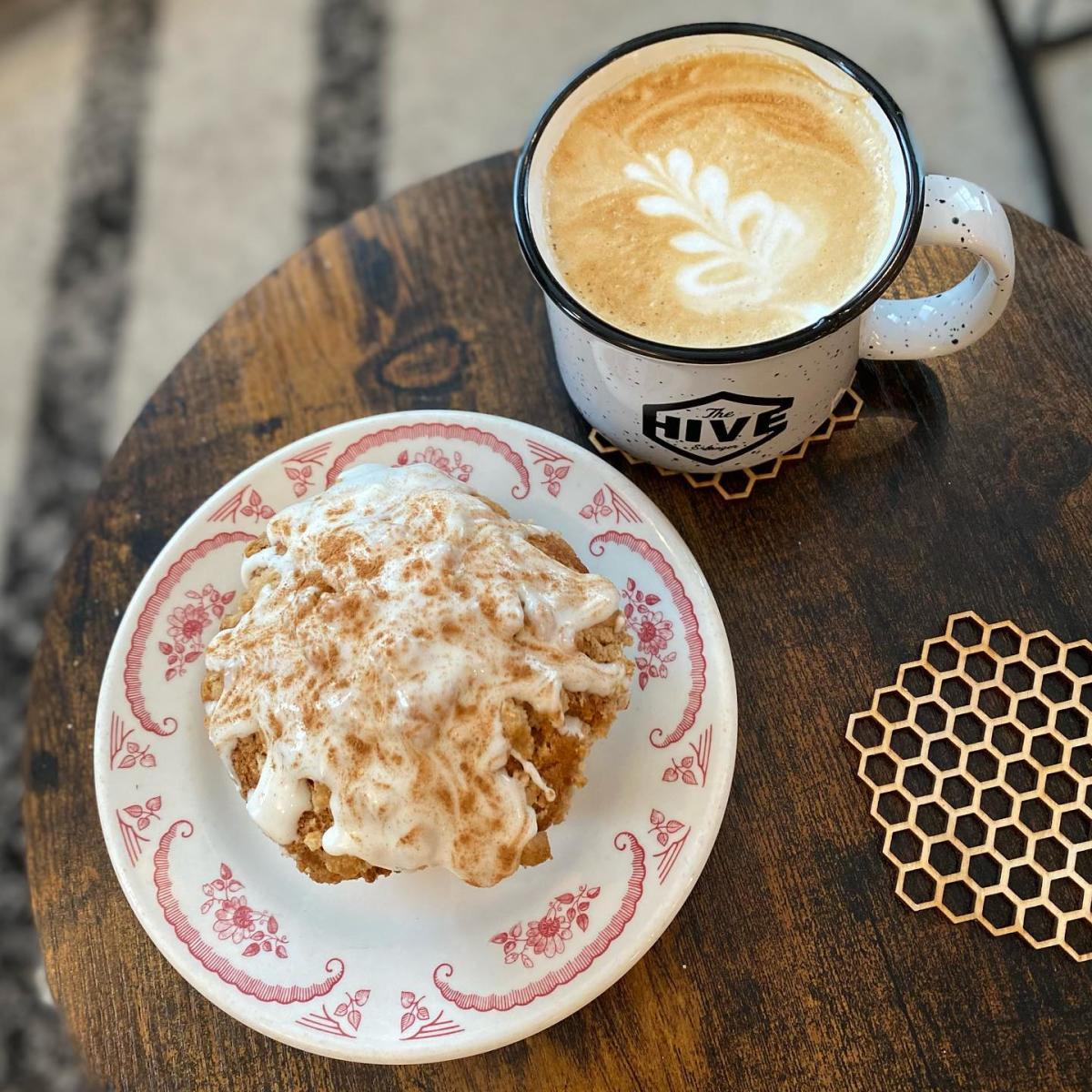 The image is an ariel view of a saucer plate with a muffin covered in icing next to a filled coffee mug on a wood table.