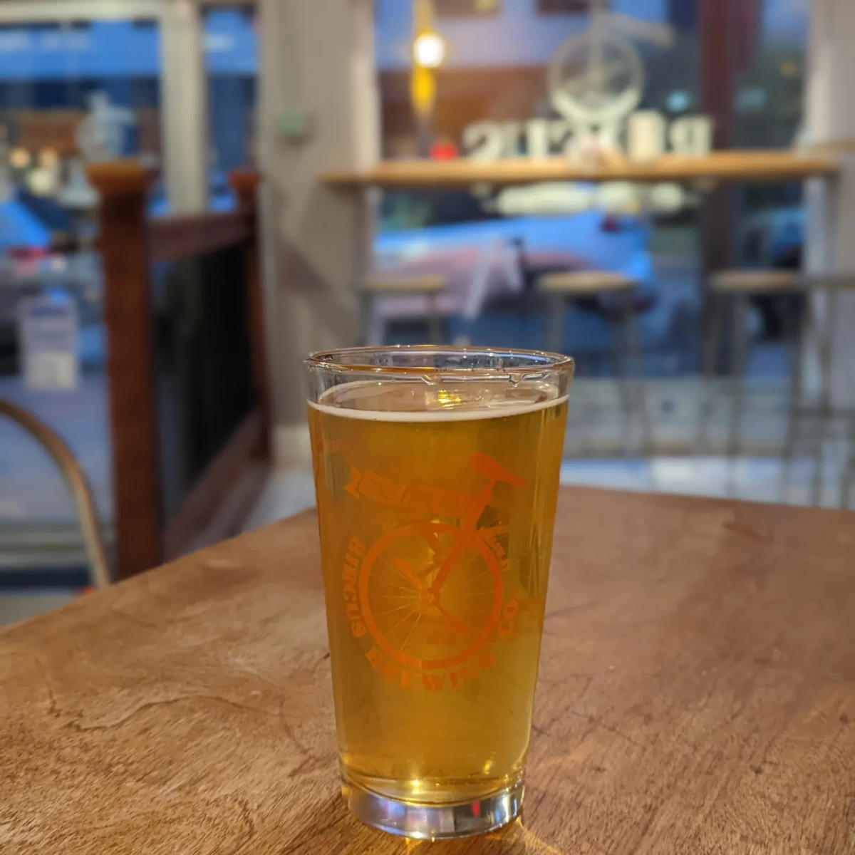 A pint glass with the Bircus Brewery logo printed on it in orange, filled with beer on a table with a view of the street outside behind it