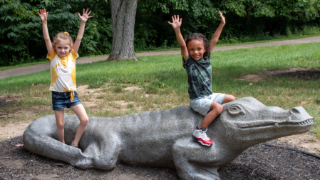 two children sitting on concrete alligator with their hands in the air, enjoying playing at Florence nature park in Florence ky