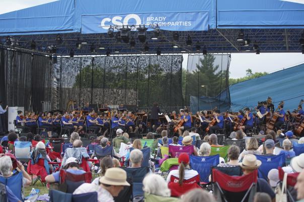 A crowd of people in pop-up lawn chairs sit by a stage and watch the Cincinnati Symphony Orchestra perform