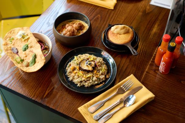 A yellow napkin with silverware sits in front of several plates on a table including a rice dish, cornbread, and a stew with hot sauce bottles to the right.