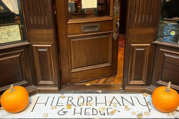 Image is of the bottom of the door and sidewalk up to the door that say's "Hierophany & Hedge" with a pumpkin on each side.