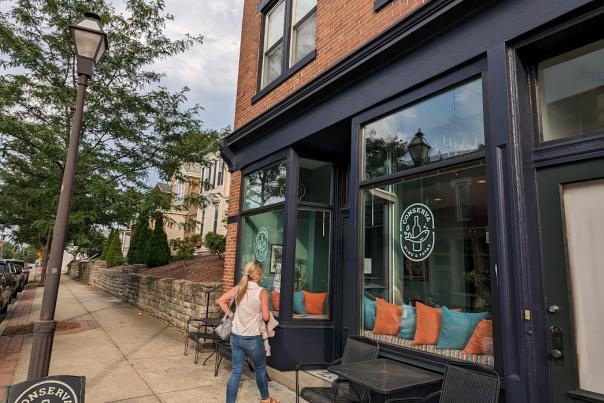 Young blonde woman entering brick building with large glass windows with sign that says Conserva Tapas