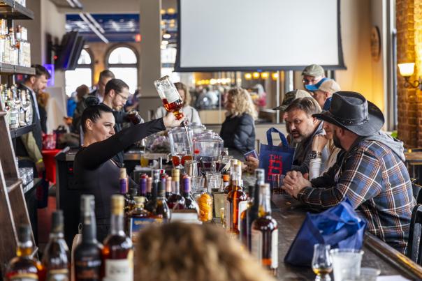 An array of bottles line the bar while a bartender pours bourbon into a glass.