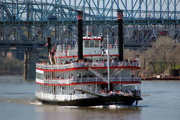 A red and white, paddlewheel riverboat cruises on the water. There are 3 decks with people standing on them.
