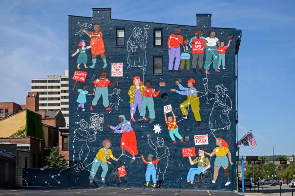 A blue background shows a variety of women, connected via constellations. They are holding signs supporting racial justice, gender equality, accessibility and healthcare for all, equal pay, and ending domestic violence.