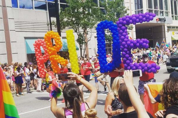 Image is of the Cincinnati Pride Parade with a group carrying the letters P.R.I.D.E made of ballons as they walk down the street.