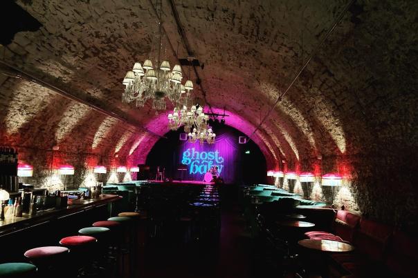A tunnel lit with white and pink lights with a sign at the end in blue reading ghost baby. Rows of barstools line the walls of the tunnel
