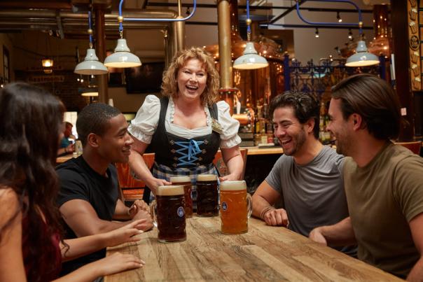 A woman in traditional German dress serving four large beers to a group at Hofbrauhaus Newport