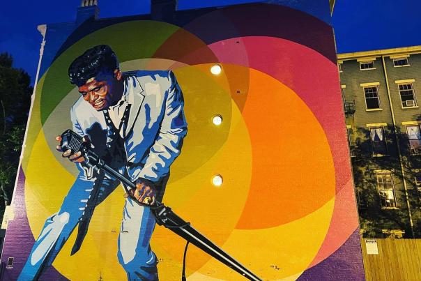 A mural of James Brown holding a microphone and leaning over to sing with yellow orange and red circles in the background